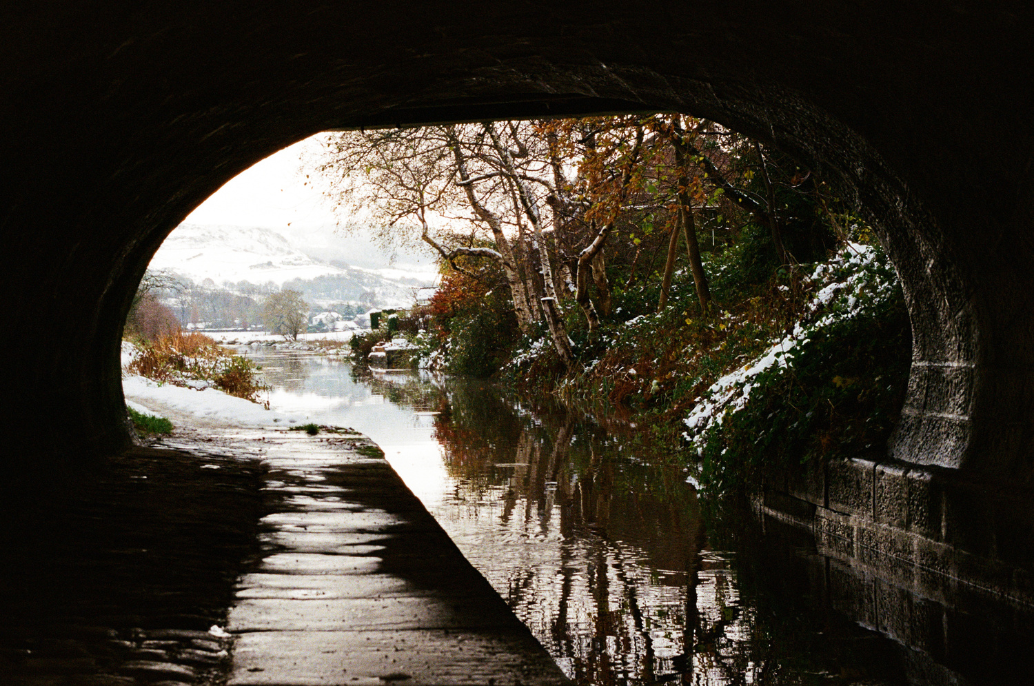 A photo looking along a canal under a bridge. The canal is slightly frozen over, and towering hills can be seen in the distance, covered in snow.