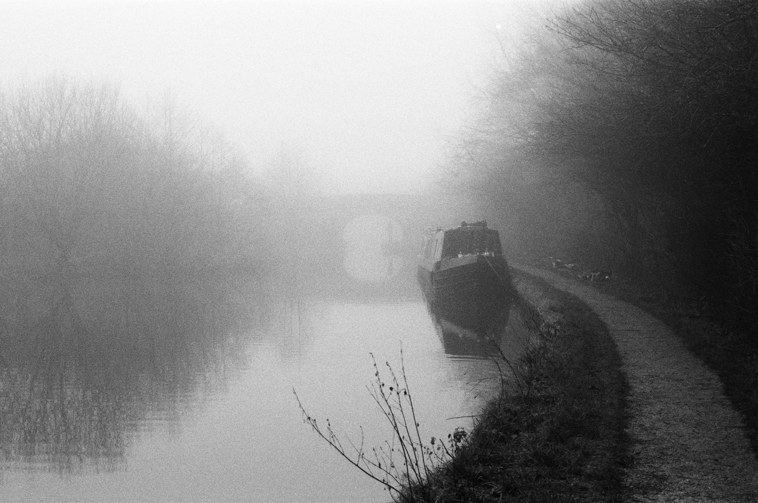 Grainy grayscale photo. A path to your right leads alongside a canal, past a narrowboat, and towards a stone bridge shrouded in dense fog. A silhouetted figure can be seen standing under the bridge - are they friend or foe?