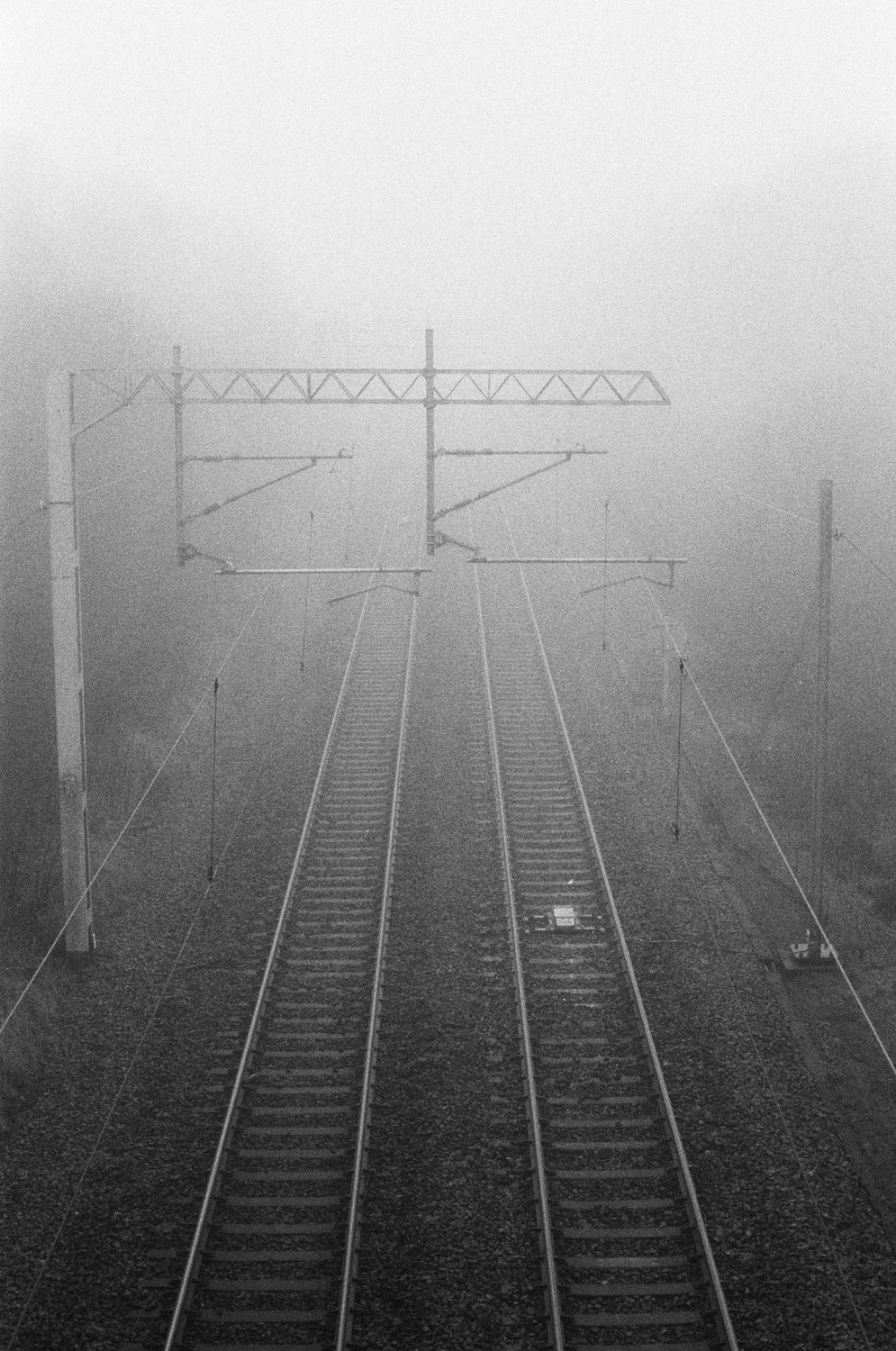 Grainy grayscale photo, looking down at two sets of rail tracks vanishing off into very dense fog.